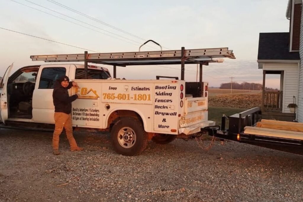 Contractor standing beside a white work truck with a ladder rack and trailer at a job site