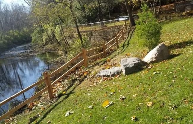 Wooden split-rail fence along a grassy creek bank with large landscaping boulders
